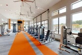 Located at 2523 Avenue M in Huntsville, Texas, this well-appointed fitness center offers a spacious, single-floor layout designed to support a full range of workout routines. The interior is immediately distinguished by its bold orange and gray carpet striping, which runs the length of the main floor and adds a vibrant, energetic aesthetic throughout the space.

The main workout area is generously proportioned, featuring a comprehensive lineup of cardio equipment including ellipticals and treadmills arranged in a row along one side. A series of large garage-style windows and clerestory windows flood the space with natural light while offering views of an outdoor pool area and surrounding grounds. High ceilings with industrial-style ceiling fans enhance airflow and contribute to an open, airy atmosphere.

Strength training is well-represented with multiple cable and resistance machines, a functional trainer, battle ropes, and a dedicated free-weight section. Full-length mirrors line portions of the walls, amplifying the sense of space and providing visual feedback during workouts.

A separate studio area at one end of the facility is outfitted for cycling and group fitness, featuring spin bikes, medicine ball racks, a wall-mounted TV, and vibrant orange accent walls. A distinct open flex zone with vaulted ceilings and additional mirrors provides space for stretching, mat work, or functional training.