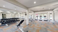 Located at 4735 Lake Trail Dr in Lisle, Illinois, this fitness center offers a well-appointed, single-floor space designed to support a comprehensive workout experience. The layout is open and spacious, framed by white structural columns that define distinct workout zones without disrupting the overall flow of the room. Patterned carpet tiles in warm orange and cool gray tones cover the floor throughout, adding visual interest while providing a comfortable surface underfoot. Recessed lighting illuminates the interior evenly, complemented by an abundance of natural light streaming through large floor-to-ceiling windows and glass doors that line the perimeter. These windows offer direct views of an outdoor pool area, creating a bright and energizing atmosphere. The cardio zone features multiple treadmills and elliptical machines positioned to take full advantage of the poolside views. A dedicated cycling area includes several stationary bikes, while a strength training section is equipped with a full dumbbell rack and adjustable benches, reflected in wall-mounted mirrors that enhance the sense of depth. A separate entry area includes built-in cubby storage for personal belongings, alongside access to an adjacent stairwell. The overall ambiance is clean, modern, and functional, making this facility a well-rounded destination for residents seeking convenient on-site fitness amenities.