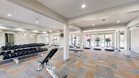 Located at 4735 Lake Trail Dr in Lisle, Illinois, this fitness center offers a well-appointed, single-floor space designed to support a comprehensive workout experience. The layout is open and spacious, framed by white structural columns that define distinct workout zones without disrupting the overall flow of the room. Patterned carpet tiles in warm orange and cool gray tones cover the floor throughout, adding visual interest while providing a comfortable surface underfoot. Recessed lighting illuminates the interior evenly, complemented by an abundance of natural light streaming through large floor-to-ceiling windows and glass doors that line the perimeter. These windows offer direct views of an outdoor pool area, creating a bright and energizing atmosphere. The cardio zone features multiple treadmills and elliptical machines positioned to take full advantage of the poolside views. A dedicated cycling area includes several stationary bikes, while a strength training section is equipped with a full dumbbell rack and adjustable benches, reflected in wall-mounted mirrors that enhance the sense of depth. A separate entry area includes built-in cubby storage for personal belongings, alongside access to an adjacent stairwell. The overall ambiance is clean, modern, and functional, making this facility a well-rounded destination for residents seeking convenient on-site fitness amenities.