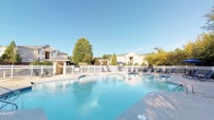 This single-level community amenity space offers a well-appointed outdoor retreat designed for relaxation and social gathering. At its centerpiece is a generously sized swimming pool with crystal-clear blue water, enclosed by a classic white picket fence that lends a clean, polished aesthetic to the entire area. The surrounding pool deck is expansive, providing ample room for sunbathing and leisure, and is furnished with multiple lounge chairs arranged to take full advantage of the open sky and natural sunlight.

Adjacent to the pool area is a covered patio that serves as an ideal outdoor gathering space. This shaded zone features a round dining table with chairs beneath a large blue market umbrella, alongside gas grills — making it well-suited for outdoor cookouts and casual entertaining. The covered structure features a pitched roof supported by columns with brick detailing at the base, adding a touch of traditional architectural charm.

The exterior of the clubhouse or amenity building is finished in warm beige siding with brick accents and a pitched gray roof, complemented by decorative wall-mounted lanterns flanking the entrance. Mature trees and thoughtfully arranged landscaping with trimmed shrubs and fresh mulch beds frame the building's entrance, enhancing curb appeal. The overall ambiance is open, bright, and inviting, with lush tree canopies providing a natural backdrop throughout the space.