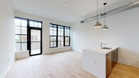 This single-floor space showcases a contemporary design aesthetic with a seamless open-concept layout connecting the living area and kitchen. Light hardwood flooring runs throughout, complementing the crisp white walls and creating a bright, airy atmosphere. Large black-framed grid windows flood the living area with natural light while offering views of the surrounding urban streetscape, and a glass door provides access to an outdoor area.

The kitchen is a standout feature, anchored by a substantial island with a white countertop, an undermount sink, and a bold slate-blue base. Dark cabinetry lines the kitchen walls, providing strong contrast against the white surfaces, while stainless steel appliances—including a gas range, microwave, and refrigerator—add a polished, functional edge. Exposed industrial ductwork and dual pendant lights overhead reinforce the modern, loft-inspired character of the space.

The bedroom benefits from its own large black-framed window, maintaining the consistent design language and filling the room with natural light. The bathroom features large-format dark tile flooring, hexagonal mosaic wall tiles in the tub-shower surround, a dark vanity, and wall-mounted lighting, delivering a clean, contemporary finish throughout.