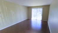 This single-floor space offers a clean, open layout designed for comfortable everyday living. The living area is a standout feature, showcasing rich, dark hardwood floors that run throughout, reflecting natural light and adding warmth to the overall aesthetic. Neutral, soft green-toned walls provide a calm and versatile backdrop, suitable for a variety of interior styles.

A large sliding glass door serves as a focal point in the main living area, flooding the space with an abundance of natural light and offering a direct view to the outdoors. Vertical blinds accompany the sliding door, providing flexible light and privacy control. The door also allows convenient outdoor access, seamlessly connecting the interior to the exterior.

The walls feature subtle textured detailing, adding a layer of visual interest without overwhelming the simplicity of the space. Clean white baseboards provide a crisp contrast against both the flooring and the wall color, contributing to a polished, well-maintained appearance.

The overall ambiance of the space is bright, airy, and move-in ready, with a straightforward layout that maximizes usability. The combination of natural light, quality flooring, and neutral finishes creates an inviting environment that is both practical and aesthetically pleasing.