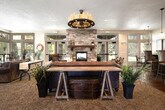 This well-appointed single-floor space offers a thoughtfully designed open-plan layout encompassing a living room, dining room, and kitchen. Polished stained concrete floors run throughout the main areas, lending a warm, cohesive aesthetic that ties the spaces together seamlessly.

The living room is a standout, anchored by a dramatic floor-to-ceiling stacked stone fireplace flanked by matching stone columns. Patterned carpet defines the seating area, while large windows and glass doors flood the room with natural light and provide views to the outdoors. Arched mirrors and eclectic furnishings add character and depth.

The kitchen is well-equipped, featuring rich dark wood cabinetry with granite countertops, a substantial island with bar seating, and pendant lighting overhead. Full-height upper cabinets maximize storage, and black appliances complement the deep-toned finishes. The kitchen flows naturally into the adjacent dining area.

The dining room is bright and inviting, with tall windows that bring in generous natural light. A chandelier overhead and a round dining table set the tone for casual yet refined gatherings.

Throughout the space, recessed lighting, stone architectural details, and carefully selected furnishings create a warm, rustic-contemporary ambiance that is both functional and visually distinctive.