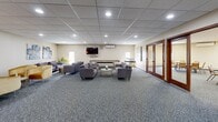 Located at 13401 Morgan Ave S in Burnsville, Minnesota, this generously sized community room offers a versatile, well-appointed space designed to accommodate a wide range of social and functional gatherings. The main hall is expansive, featuring patterned carpet flooring, a drop tile ceiling with large skylight panels and recessed lighting, and ample room for multiple round tables and chairs — making it well-suited for dining events, meetings, or community functions. A comfortable lounge area adjoins the main hall, furnished with modern gray sofas and armchairs arranged around a central coffee table, alongside a wall-mounted flat-screen television, a shuffleboard table, and a wall-unit air conditioner. The kitchenette is equipped with warm wood cabinetry, a bar-height counter with stools, a refrigerator, microwave, and water dispenser, providing convenient support for catered or self-served events. Several private rooms branch off the main space, each accessible through handsome solid wood doors with warm stained finishes. These smaller rooms feature carpeted floors, baseboard heating, and glass block windows that allow in soft natural light. The overall ambiance is clean, functional, and welcoming — a practical and comfortable community gathering space.