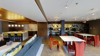 This single-floor space is a vibrant, multifunctional environment that seamlessly blends work, dining, and leisure into one cohesive layout. The open-plan design fosters a sense of connectivity throughout, with distinct zones that each serve a specific purpose while maintaining visual flow across the entire floor.

The lounge area anchors one side of the space, featuring a generous grey sectional sofa accented with patterned throw pillows. A striking red sculptural chair serves as a bold focal point, reflecting the space's energetic design language. Warm wood-paneled walls and cabinetry frame a kitchenette with a clean white countertop, adding functional convenience.

Toward the center and far end, a dining area presents white tables surrounded by bold red chairs, extending naturally into a computer workstation zone. These workstations line the back wall, which is dramatically adorned with a large-scale collage-style graphic mural featuring typography and imagery, creating an urban, editorial aesthetic.

Track lighting runs along the white ceiling throughout, providing even, directed illumination across all zones. Large-format tile flooring in a neutral tone unifies the space, while structural columns punctuate the layout. The overall ambiance is modern, edgy, and purposefully designed for communal use.
