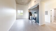 Located in Miami, FL, this one-bedroom, one-bathroom unit offers a thoughtfully designed layout that maximizes every square foot. The open living and kitchen area immediately impresses with dramatically high ceilings featuring an exposed concrete soffit, lending an industrial-chic aesthetic. Large format neutral tile flooring runs throughout, creating a cohesive and easy-to-maintain surface. A picture window fills the living area with natural light and frames green treetop and city skyline views.

The kitchen is efficiently appointed with sleek slate-blue cabinetry, white subway tile backsplash, and a full suite of stainless steel appliances including a refrigerator, range, microwave, and dishwasher — ideal for everyday cooking.

The bedroom is generously proportioned relative to the overall footprint, featuring soaring ceilings, stylish wall sconces, a large window with Miami skyline views, and a built-in shelving and closet system with double hanging rods for organized storage.

The bathroom is finished in large-format speckled tile from floor to ceiling, with a clean walk-in shower and matte black fixtures that add a modern contrast. A dedicated in-unit laundry closet with a stacked washer and dryer rounds out this well-equipped space.