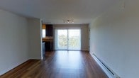 This single-floor unit offers a clean, modern layout centered around an open-concept living and dining area. Rich, dark hardwood flooring flows throughout the main living space, adding warmth and a polished aesthetic to the interior. A large sliding glass door dominates the far wall, fitted with vertical blinds and framing expansive treetop views while flooding the space with natural light. A baseboard heater runs along the wall beneath the window, providing efficient climate control. Track lighting mounted on the ceiling offers focused illumination throughout the main area. The kitchen is partially open to the living space and features dark cabinetry with a contrasting stone-style countertop, providing a sleek, contemporary look. Built-in appliances, including what appears to be a dishwasher, are integrated seamlessly into the cabinetry. The clean white walls and smooth ceilings throughout the unit create a bright, airy ambiance that complements the natural light streaming in from the balcony access point. The overall layout is functional and well-proportioned, making efficient use of the available space while maintaining an open, uncluttered feel.