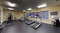 This dedicated fitness center is a comprehensively outfitted facility designed to support a wide range of workout routines. The space is finished with durable rubber flooring throughout, providing both safety and comfort during exercise. Warm yellow-toned walls and bright overhead fluorescent lighting create a clean, energizing environment. A large wall-mounted mirror spans a significant portion of the room, enhancing the sense of space and allowing users to monitor their form during workouts.

The cardio section is well-appointed with multiple treadmills and elliptical trainers from Precor, a recognized fitness equipment brand, as well as a stationary bike. Wall-mounted flat-screen televisions are positioned facing the cardio equipment, allowing users to stay entertained during their sessions. The strength training area includes a multi-station cable machine suitable for a variety of resistance exercises, a free weight rack stocked with a full range of dumbbells, and an adjustable workout bench. Stability balls in multiple sizes are also available for core training.

Entry to the fitness center is through a glass-paneled door with "FITNESS" lettering, which opens to an adjacent lounge or common area. Windows along the perimeter walls allow for natural light, contributing to an open and welcoming atmosphere throughout the space.
