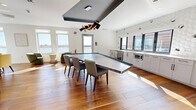 Located at 480 Morrissey Blvd in Boston's Morrissey Boulevard corridor, this thoughtfully designed lounge and amenity space offers a refined, modern environment across a single open floor. Warm hardwood flooring flows throughout, unifying the various functional zones within the room.

The space is anchored by a large, angular stone-top conference or communal table with a suspended geometric pendant light fixture above, set beneath a bold ceiling detail that adds architectural depth. Natural light pours in generously through multiple large windows, many of which frame striking waterfront views of the surrounding Boston Harbor area, creating a bright and inviting atmosphere.

A built-in kitchenette lines one wall, featuring sleek white cabinetry, white countertops, a wine cooler, microwave, and sink, all backed by a distinctive three-dimensional hexagonal tile accent wall that adds texture and visual interest.

A casual seating area includes a tufted emerald green banquette set against a detailed illustrated mural wall, accompanied by round tables and yellow upholstered chairs beneath faceted pendant lights. Additional lounge seating with curved green and yellow chairs is arranged near the waterfront-facing windows, offering a relaxed setting ideal for informal gatherings or quiet moments.