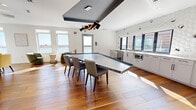 Located at 480 Morrissey Blvd in Boston's Morrissey Boulevard corridor, this thoughtfully designed lounge and amenity space offers a refined, modern environment across a single open floor. Warm hardwood flooring flows throughout, unifying the various functional zones within the room.

The space is anchored by a large, angular stone-top conference or communal table with a suspended geometric pendant light fixture above, set beneath a bold ceiling detail that adds architectural depth. Natural light pours in generously through multiple large windows, many of which frame striking waterfront views of the surrounding Boston Harbor area, creating a bright and inviting atmosphere.

A built-in kitchenette lines one wall, featuring sleek white cabinetry, white countertops, a wine cooler, microwave, and sink, all backed by a distinctive three-dimensional hexagonal tile accent wall that adds texture and visual interest.

A casual seating area includes a tufted emerald green banquette set against a detailed illustrated mural wall, accompanied by round tables and yellow upholstered chairs beneath faceted pendant lights. Additional lounge seating with curved green and yellow chairs is arranged near the waterfront-facing windows, offering a relaxed setting ideal for informal gatherings or quiet moments.