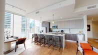This sophisticated high-rise residence is defined by its modern design and expansive floor-to-ceiling windows that frame striking city skyline views from nearly every room. The open-concept kitchen is a standout feature, showcasing two-tone wood cabinetry, sleek white quartz countertops, stainless steel appliances including a French door refrigerator and built-in microwave, and a generous island with seating for multiple guests. Pendant lighting adds warmth to the space, while warm hardwood flooring flows throughout the living areas. A cozy dining nook features a marble-top table, built-in bench seating with colorful accent pillows, and artistic wall décor, creating an intimate setting ideal for casual meals. The living room is bright and airy, anchored by large sliding glass doors that open to city views, with a neutral palette complemented by curated artwork and modern furnishings. The bedrooms are generously sized with floor-to-ceiling windows that maximize natural light and showcase the urban panorama. The bathrooms are finished with warm tile flooring, floating wood vanities, framed mirrors, and tiled shower surrounds, reflecting the same refined aesthetic found throughout the residence. Overall, this space delivers a seamless blend of contemporary style and urban living.