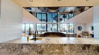 Located at 110 Constitution Dr in Menlo Park, CA, this beautifully appointed Club Room is a single-level communal gathering space designed with a contemporary, upscale aesthetic. The open-plan layout flows seamlessly across multiple distinct zones, each thoughtfully curated for both social and relaxed settings.

At the heart of the space is a striking curved bar island clad in richly veined marble, topped with a brass-finish sink and complemented by a dramatic mirrored ceiling panel above that amplifies depth and light. Adjacent to the bar, sleek blue cabinetry and bar-height seating with terracotta velvet chairs create an inviting social hub, while a wine cooler adds a refined touch.

The lounge areas showcase an eclectic yet cohesive furniture palette — including a sculptural navy curved sofa, plush rounded armchairs in warm neutrals, and bold accent seating in cobalt blue velvet. Curated artwork, decorative mirrors, and lush indoor trees in contemporary planters elevate the ambiance throughout.

Large floor-to-ceiling windows flood the interior with natural light and offer views of the outdoor terrace, enhancing the sense of openness. Light hardwood-style flooring, textured acoustic ceilings, and recessed lighting contribute to the warm, modern atmosphere. A wall-mounted display screen and a private glass-panel phone booth add functional, modern amenities to this polished communal retreat.