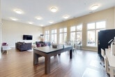 Located at 2401 Clubhouse Ct in Harrisonburg, Virginia, this single-floor clubhouse serves as a vibrant communal gathering space. The open-plan layout is anchored by soaring ceilings and an expansive wall of large, floor-to-ceiling windows that flood the interior with natural light while offering views of the surrounding community.

The main area is thoughtfully divided into multiple functional zones. A lounge area features plush gray sofas arranged around a wall-mounted flat-screen TV, complemented by a patterned area rug and accent chairs. Adjacent to this is a game zone centered around a multi-use ping-pong and billiards table with bench seating. A dining and casual seating section near the windows includes metal bistro-style chairs and glass-top tables, ideal for socializing.

Additional amenities include a computer workstation area, vending machines for snacks and beverages, and a small kitchenette nook. The interior walls are finished in a warm neutral tone, and durable wood-look flooring runs throughout, tying the space together cohesively. Recessed and flush-mount ceiling lights provide consistent, even illumination across the entire floor. A closet and entryway with interior window cutouts add architectural interest, completing this well-appointed and highly functional community clubhouse.