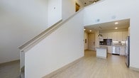 This contemporary two-story space in Dallas offers a thoughtfully designed open-concept layout that blends modern aesthetics with functional living. The main level is anchored by light hardwood flooring that flows seamlessly throughout, creating a warm and cohesive feel. Soaring double-height ceilings in the main living area amplify the sense of openness, while a sweeping staircase with a clean white profile and sleek metal railing serves as both a functional and architectural centerpiece.

The kitchen is a standout feature, appointed with light wood cabinetry, a textured stone or tile backsplash, and a granite or stone-topped island that provides generous prep and seating space. Stylish gold pendant lights hang above the island, adding a refined touch to the space. Recessed lighting throughout ensures the interior remains bright and well-illuminated.

The upper level is visible from below, offering a glimpse of an open loft-style layout complete with a ceiling fan, adding to the airy atmosphere of the home. The neutral color palette of whites and warm wood tones gives the entire space a clean, inviting ambiance well-suited to modern Dallas living.