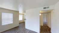 This single-floor residence offers a clean, open-concept layout designed for comfortable everyday living. The main living area is anchored by warm wood-look hardwood flooring that extends throughout, complemented by neutral beige walls and crisp white ceilings that create a bright, airy atmosphere. Multiple windows fitted with horizontal blinds allow generous natural light to fill the space, enhancing its open and welcoming feel. The living area flows seamlessly into the dining space, which is distinguished by a stylish pendant light fixture suspended overhead, adding a subtle decorative touch. A half-wall partition with a pass-through counter connects the dining area to the kitchen, providing both visual separation and convenient accessibility between the spaces. A hallway visible from the main area leads to additional rooms, with wall-mounted sconce lighting guiding the way. The consistent flooring, soft color palette, and thoughtful architectural details such as the angled ceiling transitions and defined room separations contribute to a cohesive and functional interior. The overall ambiance is calm, clean, and move-in ready, making it a versatile space suited to a variety of living arrangements.