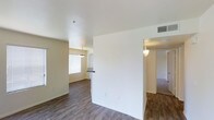 This single-floor residence offers a clean, open-concept layout designed for comfortable everyday living. The main living area is anchored by warm wood-look hardwood flooring that extends throughout, complemented by neutral beige walls and crisp white ceilings that create a bright, airy atmosphere. Multiple windows fitted with horizontal blinds allow generous natural light to fill the space, enhancing its open and welcoming feel. The living area flows seamlessly into the dining space, which is distinguished by a stylish pendant light fixture suspended overhead, adding a subtle decorative touch. A half-wall partition with a pass-through counter connects the dining area to the kitchen, providing both visual separation and convenient accessibility between the spaces. A hallway visible from the main area leads to additional rooms, with wall-mounted sconce lighting guiding the way. The consistent flooring, soft color palette, and thoughtful architectural details such as the angled ceiling transitions and defined room separations contribute to a cohesive and functional interior. The overall ambiance is calm, clean, and move-in ready, making it a versatile space suited to a variety of living arrangements.