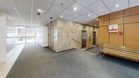 This building lobby is a thoughtfully designed communal space that serves as the primary point of entry and circulation for the building. The layout is open and efficiently organized, guiding occupants from the glass-enclosed entrance vestibule through to the elevator core and stairwell connections.

The entrance vestibule features floor-to-ceiling glass panels and automatic sliding doors, allowing natural light to filter into the lobby and providing clear sightlines to the street beyond. The flooring transitions from large-format tile near the entrance to wall-to-wall carpet throughout the main lobby area, defining distinct functional zones.

A standout architectural feature is the prominent accent wall clad in warm, veined marble-style stone, which frames the elevator bank. Two stainless steel elevator doors are set within this wall, lending a refined, professional aesthetic to the space. Adjacent to the elevators, warm wood-paneled wall sections add a complementary tone and texture.

The lobby also includes a modern parcel locker station with a touchscreen interface, offering a practical and secure package management solution for building residents. A stairwell entrance is conveniently accessible nearby. Seating in the form of a wood-and-metal bench provides a waiting area for guests.

Overhead, a recessed drop ceiling with a grid tile system houses evenly distributed recessed lighting, ensuring the lobby is well-lit throughout. Security cameras are visibly mounted, reflecting attentiveness to building safety.