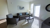 This single-floor space offers a comfortable and well-appointed living environment designed for everyday ease. The living area serves as the heart of the home, featuring soft wall-to-wall carpeting that adds warmth underfoot and a light, neutral color palette that creates a calm and inviting atmosphere throughout. Large windows allow generous amounts of natural light to pour in, illuminating the space and enhancing its airy feel. A glass-paneled door provides direct access to an outdoor balcony or patio area, further extending the sense of openness and offering a seamless connection to the outdoors. The room is thoughtfully laid out, with ample space for comfortable seating arrangements. Crown molding along the ceiling adds a refined architectural touch, lending the space a polished, finished look. The overall ambiance is clean, bright, and relaxed, making it a welcoming retreat that balances style with practicality. Neutral tones on the walls complement the natural light, creating a versatile backdrop suitable for a variety of décor styles. This space is well-suited for those seeking a comfortable, light-filled living environment with thoughtful design details throughout.