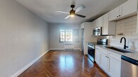 This thoughtfully designed studio offers an efficient single-floor layout comprising a main living area with an open kitchen, a hallway, and a full bathroom. The standout feature throughout the living and kitchen space is the rich, dark herringbone-patterned hardwood flooring, which lends a classic yet elevated aesthetic to the interior. A ceiling fan with an integrated light fixture anchors the main room, which is bathed in natural light from a grid-pane window and kept comfortable by a wall-mounted baseboard unit. The walls are finished in a clean, neutral light gray tone that enhances the bright, airy feel of the space. The kitchen is outfitted with white shaker-style cabinetry — both upper and lower — complemented by sleek gray countertops and a white subway tile backsplash. Stainless steel appliances include a range with oven, an over-the-range microwave, a dishwasher, and a refrigerator, alongside a matte black faucet at the undermount sink. The hallway connects to a well-appointed bathroom featuring a glass-enclosed walk-in shower with floor-to-ceiling white tile walls and a pebble-tile shower floor with matte black hardware, creating a clean, modern finish throughout.