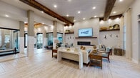 This single-floor amenity space showcases a thoughtfully designed interior that blends coastal and contemporary styles throughout. The expansive main lounge greets visitors with soaring ceilings accented by rustic exposed wood beams, light wood-look tile flooring, and recessed lighting that creates a warm, welcoming ambiance. A sleek linear fireplace anchors one wall, topped by a flat-screen display and flanked by open shelving with decorative accents, while multiple seating arrangements invite comfortable gathering.

Structural columns define distinct zones within the open floor plan, allowing natural light to flow freely from large black-framed windows that offer views to the outdoors. The beverage and lounge bar area features a dark wood island with bar seating, a dedicated coffee station, and a dramatic textured wood feature wall with an integrated entertainment screen — ideal for social gatherings. Adjacent to this, a shuffleboard table adds a recreational element.

A more intimate sitting room offers a quieter retreat, furnished with patterned armchairs, a coffee table, and a wall-mounted TV set against a decoratively paneled accent wall. Shiplap wall detailing, pendant lighting, and carefully curated décor throughout tie the space together, delivering a cohesive, upscale atmosphere suited for community engagement and relaxation.