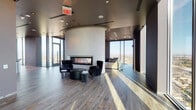 This upscale amenity lounge is a masterfully designed social space set high above the city, offering sweeping panoramic views through expansive floor-to-ceiling windows that flood the interior with natural light. The single-floor layout is thoughtfully organized into distinct zones, each with its own character and function.

The lounge area features a refined seating arrangement with plush gray sectional sofas set against a dramatic textured feature wall in dark, metallic tones, complemented by sleek leather accent chairs atop area rugs over warm wood-plank flooring. A sculptural curved column anchors a cozy fireplace seating nook, where a wide linear gas fireplace and a pair of elegant high-back chairs create an intimate retreat framed by city views.

Adjacent to this is a game and entertainment area equipped with a pool table and a wall-mounted television, lined by a counter-height bar with black swivel stools positioned directly against the glass — ideal for taking in the expansive cityscape. A dedicated bar and dining area features a richly finished wood-topped counter, pendant lighting with sleek cone fixtures, and integrated cabinetry with a display television. Throughout, the design palette of dark charcoal, warm wood tones, and metallic accents creates a cohesive, upscale ambiance suited for both relaxation and social gatherings.