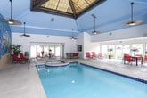 This single-story indoor pool facility offers a vibrant, resort-inspired atmosphere highlighted by a striking hand-painted tropical mural that spans an entire wall. Depicting colorful parrots, palm trees, and a coastal sunset scene, the mural sets a lively, vacation-like tone throughout the space. The generously sized swimming pool features cobalt blue tile accents and underwater lighting, while an adjacent spa provides a dedicated relaxation area. Vaulted ceilings finished in sky blue paneling contribute to the airy, open feel of the pool area, complemented by pendant lighting and a ceiling fan that promote comfortable air circulation. A glassed-in fitness area is integrated directly into the pool room, offering convenient access to exercise equipment. The surrounding pool deck is finished in a speckled terrazzo-style surface and is furnished with red lounge chairs and a dining table with matching seating, providing ample space for lounging and socializing. Large sliding glass doors and expansive windows along one side of the facility flood the lounge area with natural light and offer views of the surrounding landscape, seamlessly connecting the indoor environment to the outdoors.