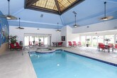 This single-story indoor pool facility offers a vibrant, resort-inspired atmosphere highlighted by a striking hand-painted tropical mural that spans an entire wall. Depicting colorful parrots, palm trees, and a coastal sunset scene, the mural sets a lively, vacation-like tone throughout the space. The generously sized swimming pool features cobalt blue tile accents and underwater lighting, while an adjacent spa provides a dedicated relaxation area. Vaulted ceilings finished in sky blue paneling contribute to the airy, open feel of the pool area, complemented by pendant lighting and a ceiling fan that promote comfortable air circulation. A glassed-in fitness area is integrated directly into the pool room, offering convenient access to exercise equipment. The surrounding pool deck is finished in a speckled terrazzo-style surface and is furnished with red lounge chairs and a dining table with matching seating, providing ample space for lounging and socializing. Large sliding glass doors and expansive windows along one side of the facility flood the lounge area with natural light and offer views of the surrounding landscape, seamlessly connecting the indoor environment to the outdoors.