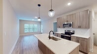 This single-floor residence showcases a thoughtfully designed open-concept layout that seamlessly connects the kitchen and living areas. The kitchen serves as a central highlight, featuring a generously sized island with a quartz countertop and an undermount sink equipped with a matte black faucet. Warm-toned, wood-grain flat-front cabinetry lines the walls and island, complemented by a classic white subway tile backsplash and a full suite of stainless steel appliances, including an electric range, microwave, refrigerator, and dishwasher. Globe pendant lights add a modern touch above the island.

The open living area extends naturally from the kitchen, bathed in natural light from large windows fitted with horizontal blinds and a glass-paneled exterior door. Light wood-look plank flooring runs throughout the main living spaces, contributing to a clean, cohesive aesthetic. The bedroom offers plush carpet flooring, a ceiling fan with light fixture, and a window providing natural light. An additional room with light flooring and recessed lighting offers flexible use.

The bathroom is well-appointed with a wide quartz vanity, wood-grain cabinetry, a large framed mirror, matte black fixtures, and a tiled tub-shower combination. Neutral tones and modern finishes unify the space throughout.