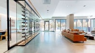 Located on Wilshire Boulevard in the heart of Los Angeles, this expansive single-floor amenity space offers a refined, contemporary design across a generously proportioned footprint. The open-concept layout is unified by polished concrete flooring that flows seamlessly throughout, lending a sleek, modern character to the entire space.

The living and lounge area is anchored by cognac leather sofas, curved sectionals, and layered area rugs, creating inviting conversation zones. A large built-in display screen with warm wood panel surround serves as a focal point, complemented by ambient lighting and recessed ceiling details. A dedicated game area with a billiard and foosball table adds a recreational dimension to the space.

A striking floor-to-ceiling glass-enclosed wine display runs along one wall, functioning as both a storage feature and a dramatic architectural statement. The hallway connecting the zones is lined with floor-to-ceiling glazed doors that open directly to an outdoor terrace, bathing the interiors in natural light and offering views of the surrounding urban landscape.

The dual-island kitchen is outfitted with stainless steel appliances, integrated wall ovens, a herringbone-patterned wood tile floor, and white quartz countertops. Warm wood cabinetry provides contrast against the crisp white finishes. Pendant lighting above each island adds refined detail, while expansive windows ensure the kitchen remains bright and connected to the outdoors.
