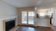This single-floor, one-bedroom, one-bathroom unit offers a well-organized layout designed for comfortable everyday living. The main living area features dark hardwood-style flooring that contrasts warmly with the neutral beige walls and textured ceiling. A classic white-mantled fireplace anchors one side of the room, adding a focal point and a touch of traditional charm. French doors with slatted blinds lead to an outdoor patio area, allowing natural light to fill the living space and providing convenient exterior access.

Adjacent to the living area, the kitchen is open and connected to the main space, allowing for an airy, communicative flow between rooms. It features light wood cabinetry, dark countertops, a black refrigerator, and a window above the sink that brings in additional natural light. Track lighting overhead ensures the kitchen remains well-lit throughout the day.

The overall ambiance of the unit is clean and neutral, with a simple, functional aesthetic. The combination of the fireplace, natural light from multiple windows, and the indoor-outdoor connection via the French doors creates a welcoming and practical living environment.