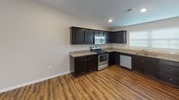 Located at 11828 Diebold Rd in Fort Wayne, Indiana, this single-story home offers a well-designed, functional layout with consistent finishes throughout. The kitchen is a standout feature, outfitted with dark cabinetry, granite-style countertops, and a full suite of stainless steel appliances including a refrigerator, electric range with microwave above, and dishwasher. Rich wood-look vinyl flooring runs through the kitchen and connecting areas, adding warmth and durability. A hallway with recessed lighting connects the main living areas to the bedrooms and bathrooms, providing a clean and organized flow. The bedrooms are carpeted for comfort, each featuring neutral walls, natural light from windows with blinds, and ceiling fans for climate control. One bedroom benefits from a large sliding glass door that floods the room with abundant natural light. The bathrooms are finished with matching dark vanities, granite-style countertops, wood-look flooring, and a tub-shower combination. One bathroom includes access to a carpeted walk-in closet with wire shelving. The overall aesthetic is contemporary and move-in ready, with a neutral color palette, textured ceilings, and clean white trim and doors throughout.