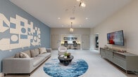 Located at 5961 SW 68th St, this single-floor multipurpose space offers a thoughtfully designed open layout with contemporary finishes and a refined aesthetic. The entrance features floor-to-ceiling glass doors framed in dark bronze, providing a welcoming transition into the interior, accented by a sculptural globe chandelier overhead and a statement potted tree beside the entry. Light-toned large-format tile flooring extends throughout, reflecting recessed lighting and contributing to the space's bright, airy atmosphere. A bold slate-blue accent wall anchors the main lounge area, which features a geometric wall art installation, a comfortable seating arrangement centered around a circular area rug in ocean-inspired tones, and a round pedestal coffee table. A recessed alcove along one wall houses a built-in workstation equipped with dual monitors, task lighting, and ergonomic seating beneath a cluster globe pendant light—ideal for a business center or collaborative workspace. On the opposite side, a wall-mounted flat-screen television sits above a sleek credenza, adjacent to a lit display niche outfitted with glass shelving showcasing decorative objects. The overall ambiance is polished and modern, balancing functional utility with an inviting, design-forward environment.