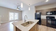 This well-appointed 1-bedroom, 1.5-bathroom residence in San Antonio, Texas spans three floors, offering a comfortable and functional layout with modern finishes throughout. The ground level features an attached garage with an automatic opener and an additional utility or storage area, providing practical everyday convenience. A staircase leads up to the main living level, where light wood-style flooring flows through a bright, open-concept space. French doors flood the living area with natural light and offer outdoor views, while a ceiling fan adds comfort. The kitchen is a standout feature, boasting a large center island with granite countertops, an undermount stainless steel sink, stylish pendant lighting, warm wood cabinetry, and black appliances including a full-size refrigerator and dishwasher. A hallway connects the kitchen to additional half-bath facilities and transitions seamlessly between rooms via light wood-tone flooring. The upper floor is home to the private bedroom, which is generously sized with plush wall-to-wall carpeting, a ceiling fan, and its own set of French doors that invite natural light. The overall aesthetic is clean and contemporary, with a neutral color palette of soft beiges and whites creating a calm, welcoming ambiance throughout the home.