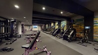 Located in Watertown, MA, this fitness center offers a comprehensive, single-floor layout designed to accommodate a wide range of workout needs. The space features a dark, modern aesthetic with charcoal-toned walls, black-beamed ceilings, and durable rubber flooring throughout, creating a focused and energetic atmosphere.

The cardio zone is lined with a row of treadmills, stair climbers, and elliptical machines, each equipped with individual screens for on-demand entertainment. Vibrant, color-coded wall graphics run along the length of this area, adding a bold visual element to the space. Mounted flat-screen televisions provide additional viewing options for members during workouts.

The strength training area includes a multi-station cable machine, a full dumbbell rack with benches, and a variety of adjustable equipment for targeted muscle work. Kettlebells and exercise mats are readily available for floor-based training. A dedicated functional training zone offers open floor space with medicine balls, spin bikes, and a wooden cubby storage unit, ideal for group or circuit-style workouts.

Structural concrete columns accent the interior, complementing the industrial design. Recessed lighting provides consistent illumination throughout. Additional utility spaces, including an A/V closet, support the facility's operational needs. The overall layout is efficient, organized, and designed to support diverse fitness routines.