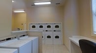 Located at 111 Coopers Ridge Blvd in Ladson, South Carolina, this single-floor communal laundry facility is designed to accommodate residents with efficiency and convenience. The room features a clean, neutral palette with warm beige walls and light-toned tile flooring that contribute to a bright and welcoming atmosphere. Overhead fluorescent lighting ensures the space is well-illuminated throughout. The facility is equipped with multiple top-load washing machines and a bank of stacked front-load dryers arranged along the back wall, providing ample laundry capacity for simultaneous use. Folding counters are positioned conveniently within the space, offering residents a practical surface for sorting and folding laundry. The layout is thoughtfully organized to allow easy movement between machines and work surfaces. Ventilation is provided through ceiling vents, maintaining air circulation within the room. The overall design prioritizes functionality and ease of use, making this laundry facility a practical and well-maintained communal amenity for the residents of Coopers Ridge.