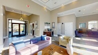 Located at 2615 Mt Zion Pkwy in Jonesboro, GA, this single-floor community space offers a well-appointed layout comprising a spacious living room and a connecting hallway. The living room immediately impresses with its soaring ceilings and a coffered tray ceiling detail, lending the space a grand, open feel. Large arched windows and floor-to-ceiling glass panels along with French doors flood the interior with natural light, offering views of an outdoor deck area. The neutral warm-beige walls provide a welcoming backdrop for the contemporary furnishings, including plush sofas, accent chairs, and a patterned multi-tone carpet that adds visual interest throughout. A built-in beverage and refreshment station with dark wood cabinetry serves as a practical amenity within the living area. A dedicated workstation nook features a built-in desk, multiple computer terminals, and additional seating, making it a functional space for business needs. Recessed lighting and a modern geometric pendant fixture enhance the overall ambiance. The hallway connects seamlessly to the living room, maintaining the same refined aesthetic. The combination of high ceilings, plentiful natural light, and thoughtful furnishings creates a bright, comfortable, and highly functional communal environment.