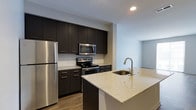 Located at 3443 Park Street in Grove City, Ohio, this single-floor residence offers a clean, modern aesthetic throughout. The kitchen serves as a standout feature of the space, designed with a cohesive contemporary style that balances function and visual appeal. Rich, dark espresso-toned cabinetry lines the walls, providing generous storage from floor to ceiling, while stainless steel appliances — including a refrigerator, electric range, and over-the-range microwave — lend a polished, professional finish. The countertops throughout the kitchen are finished in light speckled granite, offering a striking contrast against the dark cabinetry. A central island extends the workspace and features a single-basin undermount sink with a modern arc faucet, making it an ideal hub for both meal preparation and casual gatherings. Wood-look flooring runs throughout, adding warmth and continuity to the space. Recessed lighting illuminates the kitchen efficiently, contributing to the bright and open atmosphere. The kitchen opens toward an adjacent living area, creating a connected, flowing layout well-suited for everyday living. The overall design reflects a thoughtful blend of modern materials and practical functionality.