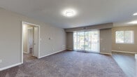 This single-floor residence presents a well-organized layout designed for comfortable everyday living. The living area is generously proportioned, finished with neutral-toned carpet and painted walls in warm gray. A large sliding glass door fitted with vertical blinds fills the space with natural light and provides direct access to an outdoor patio, offering views of surrounding greenery. The open layout connects seamlessly to the kitchen, which has been updated with crisp white shaker-style cabinetry, brushed nickel hardware, marble-look countertops, a white subway tile backsplash, and stainless steel appliances including a range with overhead ventilation and a refrigerator. Light wood-look flooring completes the kitchen's clean aesthetic. The bedrooms are carpeted and feature neutral walls, windows with blinds, and built-in closets for storage. The primary bedroom includes direct access to a private en-suite bathroom finished with wood-look tile flooring, a tub-shower combination, and a toilet. A second bathroom features a white vanity with a single sink, a large mirror, and brushed nickel fixtures. Throughout the unit, the design maintains a cohesive, understated palette that creates a calm and move-in-ready atmosphere.
