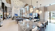 Located at 425 Centerpointe Circle in Altamonte Springs, FL, this upscale amenity lounge is a masterfully designed communal space that blends modern aesthetics with resort-style recreation. The space is defined by dramatic double-height ceilings that amplify the sense of openness, complemented by an abundance of natural light streaming through floor-to-ceiling windows dressed in sheer drapery, offering views of the surrounding outdoor pool area and landscaping.

The interior design is anchored by polished large-format tile flooring and a striking veined marble island with white upholstered bar seating, serving as both a functional centerpiece and a design statement. A cascading sculptural pendant light installation overhead adds a gallery-worthy focal point. The lounge area features a bold textured accent wall with an integrated fireplace and wall-mounted television, surrounded by curated seating arrangements ideal for socializing.

The game room component includes full-size billiards tables, a shuffleboard table, and arcade gaming stations, catering to a wide range of recreational interests. A built-in bar and kitchen area with stainless steel appliances and open shelving provides convenient refreshment access. Warm wood slat ceiling details, designer globe pendant lighting, and a carefully curated mix of furnishings throughout contribute to the space's polished, contemporary ambiance.