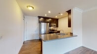 Located at 6631 Tanager Lane in Hilliard, Ohio, this single-floor residence offers a well-designed open-concept layout that connects the kitchen and living areas seamlessly. The kitchen is a functional centerpiece, featuring dark espresso cabinetry with ample storage, a peninsula with a double-basin sink and gray countertops, and a full suite of stainless steel appliances including a refrigerator, range, microwave, and dishwasher. Warm hardwood-style flooring runs throughout the main living and kitchen areas, complementing the clean white walls and crown molding detail. The living area is bright and open, with a door leading to an outdoor space and a large window that invites natural light and outdoor views. The bedroom is carpeted for comfort, with a generously sized window providing additional natural light. The bathroom is finished with classic black-and-white mosaic tile flooring, a tub/shower combo, a dark-cabineted vanity with a white countertop, and a large mirror. A dedicated in-unit laundry closet houses a stacked full-size washer and dryer, adding everyday convenience. The overall aesthetic is modern and neutral, offering a clean and move-in-ready environment.