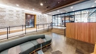 This single-floor space serves as the amenity and lobby level of a modern residential building, combining functionality with refined contemporary design throughout. The entrance area welcomes residents and guests with polished concrete flooring, a textured decorative wall feature with subtle backlighting, and a curved green sofa that adds a warm, inviting touch. Floor-to-ceiling glass partitions allow natural light to flow freely, while black steel railings and raised platform areas define distinct zones within the open layout.

A dedicated conference and collaboration room features a large blue communal table surrounded by wooden chairs, built-in shelving stocked with books, an integrated display screen, and curated artwork on the walls — creating a productive yet comfortable environment. Adjacent to this area, a generously sized mail and package room is outfitted with sleek, numbered dark metal mailboxes arranged across island units and wall-mounted rows, all framed by warm wood trim and illuminated by elegant pendant lighting and recessed fixtures.

The elevator lobby showcases a dramatic textured dark tile accent wall with integrated lighting, polished stainless steel elevator doors, and greenery that softens the industrial aesthetic. Throughout the space, the consistent use of concrete floors, warm wood tones, and carefully placed lighting creates a cohesive, upscale ambiance that balances practicality with modern sophistication.