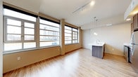 This single-floor space showcases a thoughtfully designed open-concept layout that seamlessly connects the living area and kitchen. Large, floor-to-ceiling windows line the exterior wall, flooding the interior with abundant natural light while offering sweeping views of the surrounding urban skyline. Rounded structural columns frame the window wall, adding an architectural character to the space.

The flooring throughout is a light-toned hardwood-style material that enhances the bright, airy atmosphere. A track lighting system runs along the ceiling, providing flexible, adjustable illumination across the main living area.

The kitchen area features a sleek island with a white countertop and a dark wood-toned base, complemented by an undermount sink and modern faucet. Two pendant lights hang above the island, adding a warm, focused glow that contrasts elegantly with the cooler tones of the surrounding space.

The overall ambiance is contemporary and urban, with a neutral color palette of warm beige walls and white ceilings that creates a versatile, welcoming environment well-suited to a variety of uses.