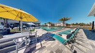 Located at 1600 LPGA Blvd in Daytona Beach, Florida, this resort-style outdoor amenity space is designed to deliver a refined, leisure-focused experience across two levels. At the heart of the space is a generously sized, crystal-clear swimming pool with a smooth tile finish, surrounded by an expansive paver deck finished in a herringbone pattern. Rows of teal lounge chairs line the poolside, complemented by large yellow market umbrellas that provide shaded retreat throughout the day. Lush palm trees are strategically placed throughout the landscape, reinforcing the tropical Florida ambiance under open blue skies. A covered outdoor lounge area adjacent to the clubhouse building features comfortable modular seating arrangements, ceiling fans, and direct views of the pool and surrounding grounds. The clubhouse structure itself is architecturally distinguished by soaring floor-to-ceiling glass curtain walls that flood the interior with natural light and create a seamless visual connection between indoor and outdoor spaces. Stone steps with metal railings lead to the patio, accented by contemporary bowl planters and lattice lounge chairs. The multi-story residential building visible in the background anchors the community setting, completing the cohesive, upscale aesthetic of this well-appointed outdoor gathering destination.