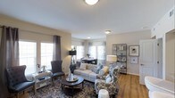 This single-floor residence offers a thoughtfully designed living space that balances comfort and elegance. The open-concept layout seamlessly connects the living and dining areas, creating a cohesive and inviting atmosphere throughout.

The living area is anchored by a tufted sofa paired with patterned accent chairs, centered around a dark, circular coffee table atop a richly detailed area rug. A stylish open shelving unit along one wall provides both display and storage space, while a combination of floor lamps and flush-mount ceiling fixtures ensures warm, layered lighting throughout.

Large windows dressed with sheer and grommet-panel curtains line the walls, allowing abundant natural light to fill the room and highlight the warm-toned hardwood flooring underfoot. The neutral wall palette in soft beige tones complements the varied textures and patterns of the furnishings, lending the space a refined yet approachable character.

Adjacent to the living area, the dining space accommodates a round table with seating for several guests, positioned near bay-style windows that further enhance the sense of openness. Clean white ceilings and smooth wall finishes contribute to the bright, airy ambiance that defines this well-appointed residence.