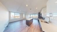 This contemporary single-floor residence showcases a thoughtfully designed open-concept layout that seamlessly connects the kitchen and living areas. Warm hardwood flooring runs throughout the space, complementing the clean, modern aesthetic. Tray ceilings with recessed lighting add architectural interest and an elevated sense of height to the main living area.

The kitchen is a standout feature, equipped with stainless steel appliances including a gas range, microwave, dishwasher, and refrigerator. Two-toned cabinetry pairs dark lower cabinets with sleek, high-gloss white upper cabinets, accented by a classic subway tile backsplash and light quartz countertops. A freestanding island provides additional prep space and storage, anchoring the kitchen within the open floor plan.

The expansive living area is bathed in natural light, courtesy of large, multi-pane windows that wrap around the space and frame panoramic views of the surrounding landscape and skyline. The generous proportions of this area offer flexible arrangements for furnishing.

The bedroom features a tray ceiling with recessed lighting, hardwood floors, and ample closet space provided by two sets of double doors, offering excellent storage. The overall ambiance of the space is bright, airy, and modern, with a neutral palette that provides a versatile backdrop throughout.