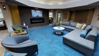 This single-floor lounge space offers a thoughtfully designed interior that blends modern aesthetics with comfortable functionality. The room immediately draws attention with its striking vibrant blue carpet, which anchors the seating area and adds a bold pop of color against the more neutral tones of the surrounding décor.

The focal wall features a sleek, dark wood-paneled surround housing a linear electric fireplace below a large wall-mounted flat-screen television, creating an entertainment hub that serves as the room's centerpiece. This combination provides both warmth and visual interest for occupants.

The seating arrangement is generous and well-considered, with a large L-shaped gray sectional sofa paired with accent armchairs and a round white marble-topped coffee table with a metallic base. The layout encourages conversation and relaxed gathering.

Natural light filters in through windows visible in the background, offering views of surrounding greenery and brightening the space. A recessed ceiling detail adds architectural depth overhead, while the overall color palette — cool blues, warm grays, and dark wood tones — creates a cohesive and inviting ambiance. Decorative accessories including vases and floral arrangements add finishing touches throughout. This space is well-suited for community lounging, informal meetings, or social gatherings.
