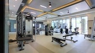 This contemporary fitness center offers a thoughtfully designed workout environment suited for a wide range of exercise routines. The space is anchored by a striking coffered ceiling with recessed linear lighting and warm ambient LED accents, giving the room a polished, upscale aesthetic. Ceiling fans are integrated throughout, ensuring comfortable air circulation during workouts.

The facility is generously outfitted with a comprehensive selection of equipment. A Synergy360 functional training system anchors the strength training area, complemented by weight benches and a variety of resistance machines. The cardio zone, positioned along the perimeter near the large floor-to-ceiling windows, features treadmills, elliptical trainers, and upright bikes, allowing users to exercise while enjoying views of the surrounding outdoor area.

The flooring throughout is a durable, light-toned material designed for both comfort and easy maintenance. Natural light pours in through the expansive windows along one wall, creating a bright and energizing atmosphere. The overall layout is open and spacious, providing ample room between equipment stations to move freely and safely. The design balances functionality with a high-end aesthetic, making this fitness center a standout amenity.