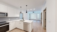 This single-floor residence showcases a clean, contemporary design throughout, characterized by a bright white palette, light wood-look flooring, and modern matte black hardware accents. The open-concept kitchen is a standout feature, anchored by a large center island with a white quartz countertop, an undermount sink, and a stainless steel dishwasher. Sleek flat-front white cabinetry lines the walls from floor to ceiling, complemented by a gray subway tile backsplash. Stainless steel appliances — including a side-by-side refrigerator with ice and water dispenser, an electric range, and an over-the-range microwave — complete the well-appointed kitchen. Striking black pendant lights hang above the island, adding a bold design contrast. The kitchen flows seamlessly into a spacious living area filled with natural light from large windows fitted with white blinds and a ceiling fan overhead. The bedroom features a stylish sliding barn door that opens to a walk-in closet, with a ceiling fan adding comfort. The bathrooms are finished in crisp white tones — one includes a glass-enclosed shower with a long vanity, while another offers a soaking tub with white subway tile surround and built-in shelving. An in-unit stacked washer and dryer adds everyday convenience to this thoughtfully designed space.