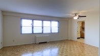 This single-floor residence offers a straightforward and functional layout with timeless design details. The main living area is immediately welcoming, characterized by its classic parquet wood flooring arranged in a distinctive block pattern that adds warmth and texture underfoot. A row of large windows fitted with horizontal blinds lines one wall, allowing abundant natural light to fill the room and creating a bright, airy atmosphere. A ceiling fan is centrally positioned overhead, contributing to both comfort and air circulation throughout the space. A baseboard heating unit sits beneath the windows, ensuring year-round climate control. The neutral white walls and smooth ceiling provide a clean, versatile backdrop that easily accommodates a variety of décor styles. A doorway visible at the far end of the living area leads to additional rooms, suggesting a practical and connected floor plan. Overall, the space presents a well-lit, efficiently designed interior with classic flooring and ample natural light as its standout features.