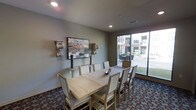 This single-floor communal space is designed to offer a comfortable and functional environment for gatherings and meetings. The dining area serves as the focal point, featuring a large rectangular light wood table surrounded by upholstered chairs with nailhead trim detailing, providing seating for a sizeable group. The room is finished with neutral gray walls that create a calm, welcoming atmosphere, complemented by recessed lighting that ensures a bright and inviting interior. Decorative wall art and ornamental corbel brackets add character and a touch of refined style to the space. Large floor-to-ceiling windows and a sliding glass door along one wall flood the room with natural light while offering direct views of an outdoor patio area, where additional seating and landscaping can be seen. The patterned carpet flooring adds visual interest and warmth underfoot. The overall ambiance balances practicality with thoughtful design, making it well-suited for both casual social gatherings and more structured events. The seamless connection between the interior dining area and the outdoor space enhances the sense of openness and livability throughout.